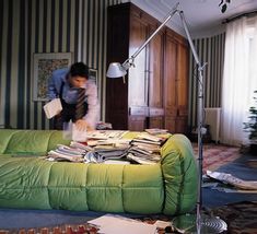 a man standing next to a green couch in a living room filled with books and papers