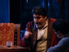 two men sitting at a table talking to each other in front of a red chair