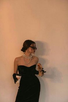 a woman in a black dress and pearls is leaning against a wall with her hands on her hips