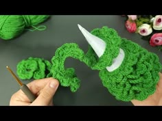 someone crocheting a green hat on top of a table with yarn and flowers