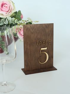 a wooden table number sitting next to a wine glass and vase with pink roses in it