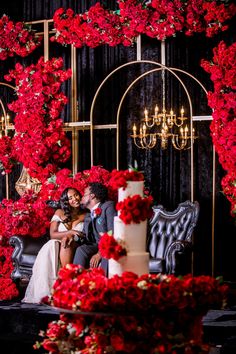 a bride and groom sitting in front of a wedding cake with red flowers on it