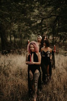 three women in bodysuits standing in tall grass with trees in the back ground