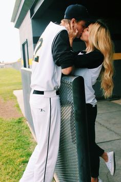 a man and woman kissing on the side of a baseball dugout with grass in the background