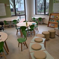 an empty classroom with green chairs and round tables in front of two large windows that look out onto the park