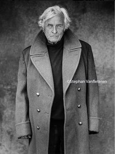 an old man wearing a coat and standing in front of a wall