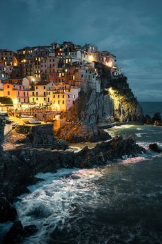 the village is lit up at night on top of a cliff by the ocean with waves crashing in front of it