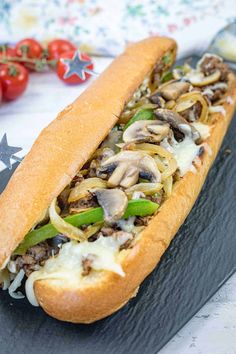 a long sandwich with mushrooms, peppers and cheese on a black plate next to tomatoes