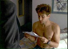 a shirtless man reading a paper while standing in front of another person on a bed