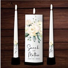 two white candles with flowers on them sitting next to each other in front of a wooden wall