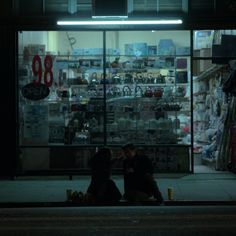 two people sitting on the sidewalk in front of a store at night with their backs to each other