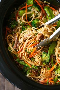 someone using tongs to stir noodles in a crock pot with broccoli and carrots