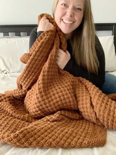 a woman laying on top of a bed covered in a brown knitted blanket and smiling at the camera