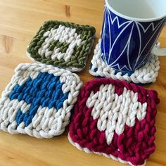 three crocheted coasters sitting on top of a wooden table next to a coffee cup