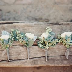 four boutonnieres with white flowers and greenery are arranged on a piece of wood