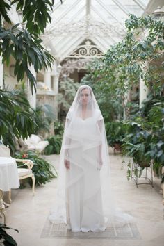 a woman in a white wedding dress and veil standing on a rug with greenery behind her