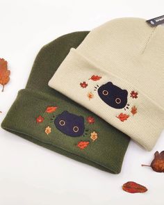 two beanies with cats on them sitting next to leaves