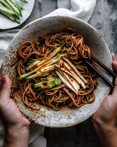 two hands holding a bowl of noodles with chopsticks