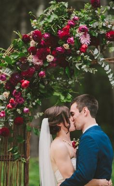 a bride and groom are kissing under an arch with flowers on the top, surrounded by greenery