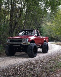 a red pick up truck driving down a dirt road in the woods with trees on both sides