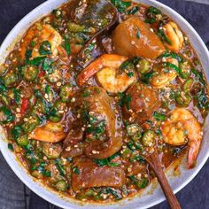 a white bowl filled with shrimp and vegetable stew next to a wooden spoon on top of a table