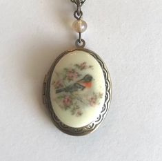 Here is a pretty bird locket necklace. This necklace features an oval shaped brass locket with a vintage style cameo cabochon in the center. The cabochon depicts a sweet spring robin bird perched on a cherry blossom branch, the cameo is made from resin. This locket has an engraved floral pattern along the front side border and it is suspended from a glass bead. Locket comes on a sturdy soldered brass chain that is closed with a solid brass clasp. Perfect for spring or summer. All jewelry comes i Bird Locket, Robin Necklace, Locket Vintage, Side Border, Victorian Locket, Blossom Branch, Cherry Blossom Branch, Robin Bird, Bird Gifts