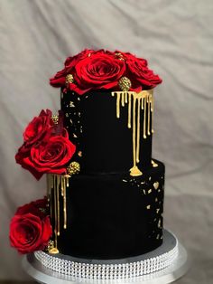 a three tiered black and gold cake with red roses on the top, drizzled in gold