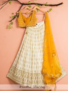 A three-piece ivory and yellow mirror lehenga set from the Anisha Shetty collection. This gorgeous ivory georgette lehenga is paired with a yellow mirror blouse in raw silk fabric. The ivory lehenga is enhanced with heavy sequence work all over with scalloped edging. The V neck mirror blouse has scalloped edging all over with a ghungroo tassel tie-up at the back.  This outfit is completed with a yellow net dupatta with heavy embroidery scalloped edging. White Bollywood Choli In Chinon, Designer White Chinon Choli, White Chinon Choli For Designer Wear, Designer Wear White Choli In Chinon, Cream Georgette Lehenga For Festivals, White Chinon Lehenga For Navratri, White Chinon Lehenga For Festive Season, White Chinon Lehenga For Reception, Yellow Dola Silk Choli With Mirror Work