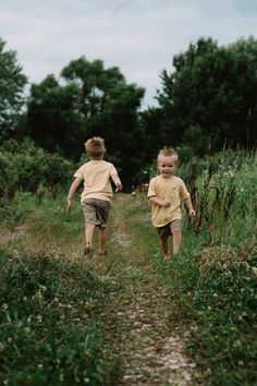 two young boys running down a dirt path in the middle of tall grass and trees