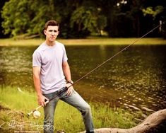 a young man standing on a log with a fishing rod in his hand while posing for the camera