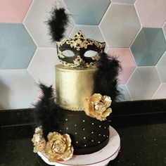 a three tiered cake decorated with black and gold feathers, flowers and a masquerade