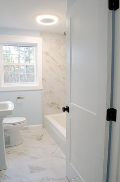 a white bathroom with marble flooring and walls, along with a window above the toilet