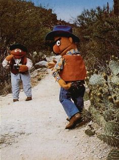 two stuffed animals walking down a dirt road next to cactus bushes and trees on either side of them