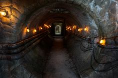 an underground tunnel with lights on the side and stone walls, leading to another tunnel