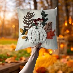 a hand holding up a decorative pillow with leaves and pumpkins on it in the fall