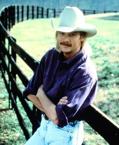 a man in a cowboy hat leaning on a fence with his arms crossed and looking at the camera