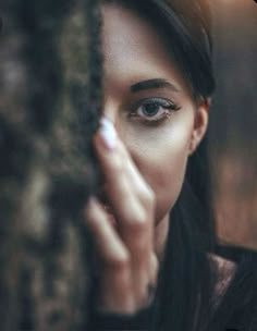 a woman with her hand on her face looking out from behind a tree trunk, while the background is blurry