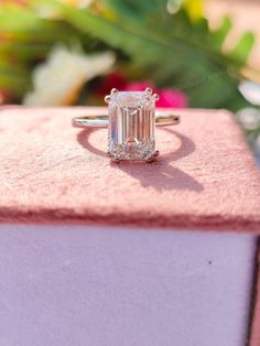 an emerald cut diamond ring sitting on top of a pink velvet box with flowers in the background