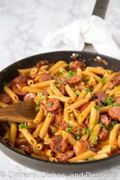 a skillet filled with pasta and sausage on top of a white tablecloth next to a wooden spoon