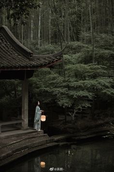 a woman standing in front of a pond with a lantern under her arm and trees behind her