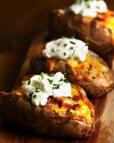 three baked potatoes topped with sour cream and chives on a wooden cutting board, ready to be eaten