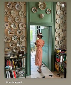 a woman in a dress and hat is looking at plates on the wall behind her