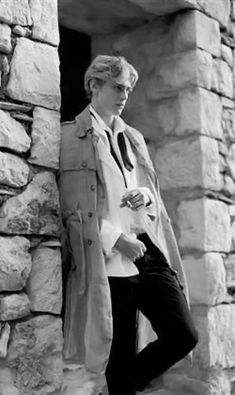 black and white photograph of a man in trench coat leaning against stone wall with hand on hip