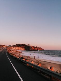 an empty highway next to the ocean with people walking on the beach in the distance