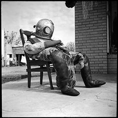 a man sitting on top of a wooden chair wearing a diving suit and boots next to a brick wall