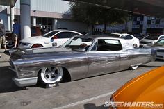 an old silver car parked next to other cars in a parking lot with people walking by