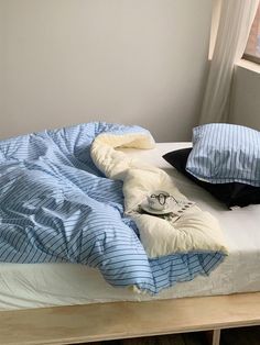 an unmade bed with blue and white comforters on it, next to a window