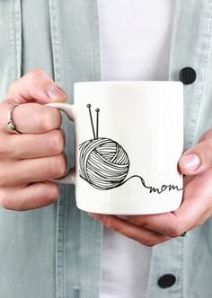 a woman holding a coffee mug with a ball of yarn on it and the word mom written in black ink