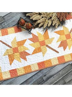 an orange and yellow quilted table runner with autumn leaves on it next to a pine cone