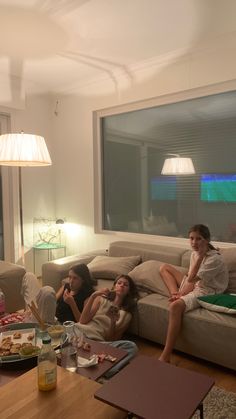 three women sitting on a couch in a living room with food and drinks all around